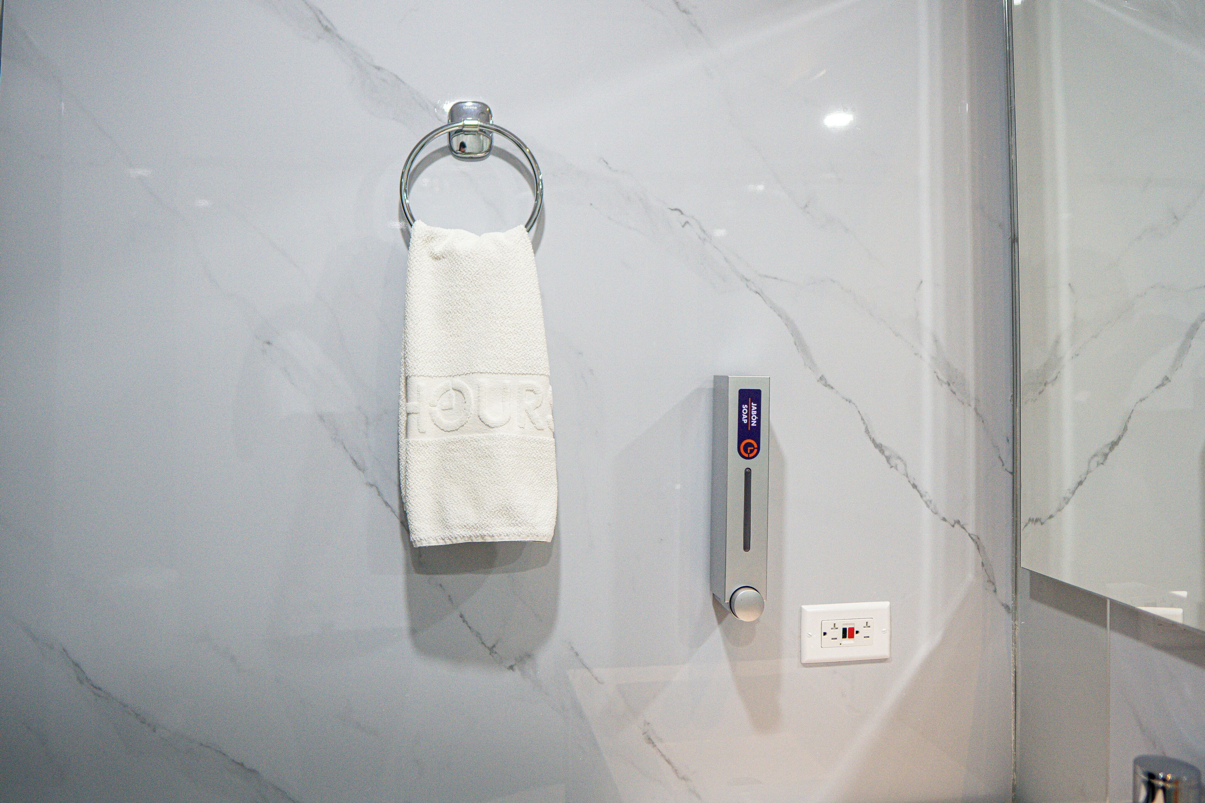 A white towel hanging on a metal ring against a marble wall, a soap dispenser, and a power outlet.