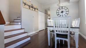 Un coin repas avec une table et des chaises blanches, une horloge au mur et un escalier menant à une autre pièce.