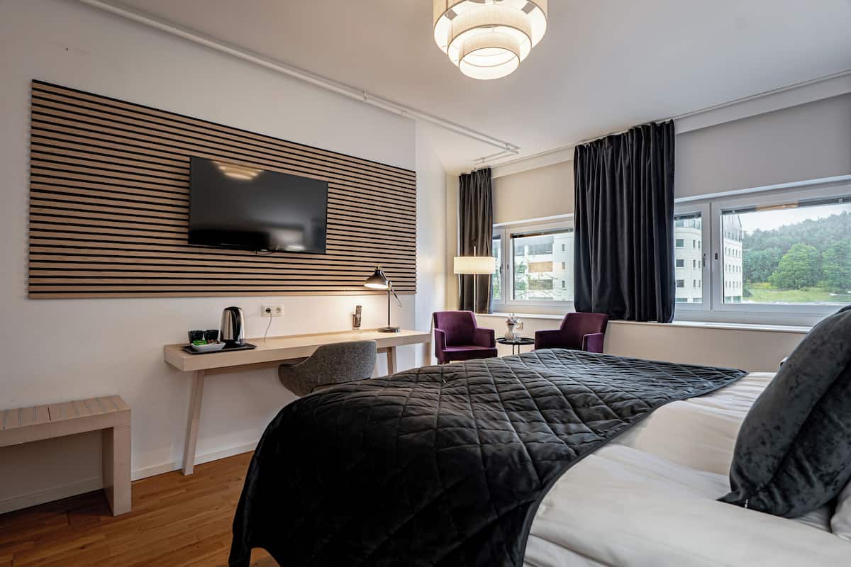 Ein modernes Hotelzimmer mit Bett, Schreibtisch, Sessel, Fernseher und einem fensterbepflanzten Fenster.
