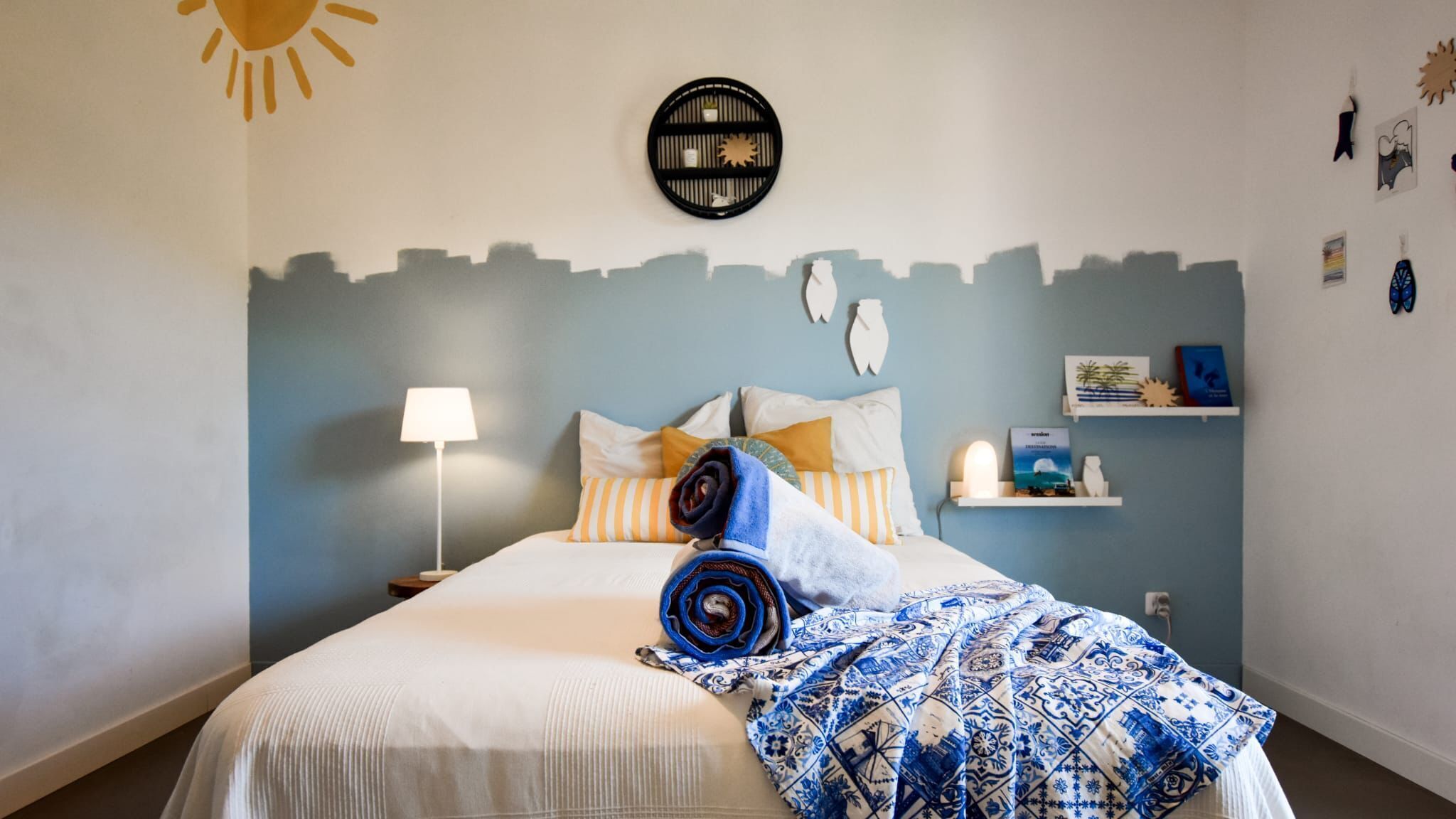 Una cama bien hecha con un edredón estampado, una lámpara de noche y una estantería de pared con libros y objetos decorativos.