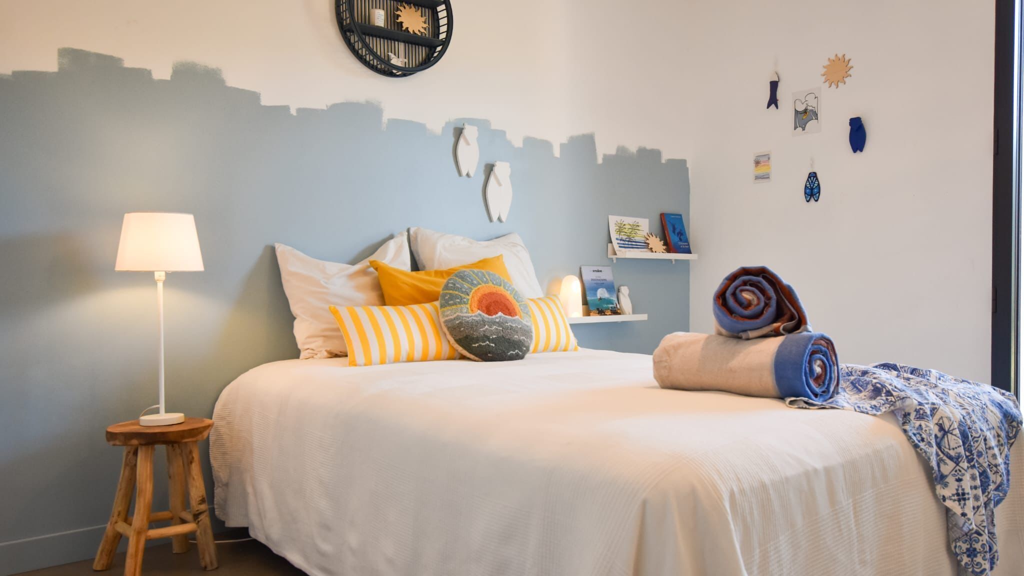 A neatly made bed with pillows and a rolled towel, a bedside lamp, and a small wooden stool.