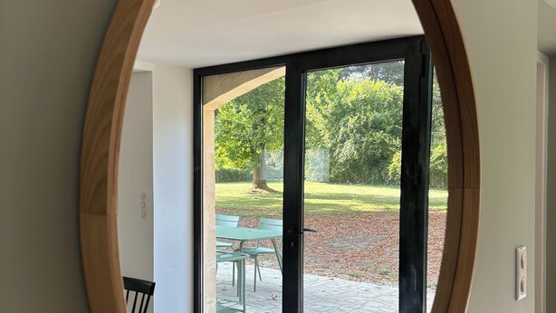 A room with a large oval mirror reflecting a patio area with a table and chairs, a tree, and a lawn. - Superb barn in the Dordogne, in the heart of Trémolat (Trémolat)