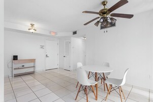 A spacious room with a white ceiling fan, a white table with two chairs, a wooden desk, and a white door with an exit sign.