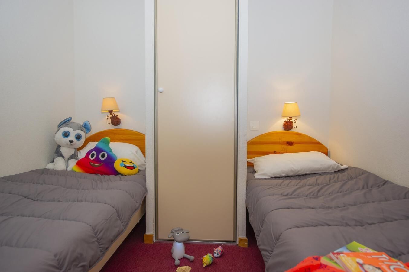 Ein Hotelzimmer mit zwei Einzelbetten, jeweils mit einem hölzernen Kopfteil und einer Lampe. Auf den Betten liegen kuschelige Stofftiere und auf dem Boden ein kleines Spielzeug.