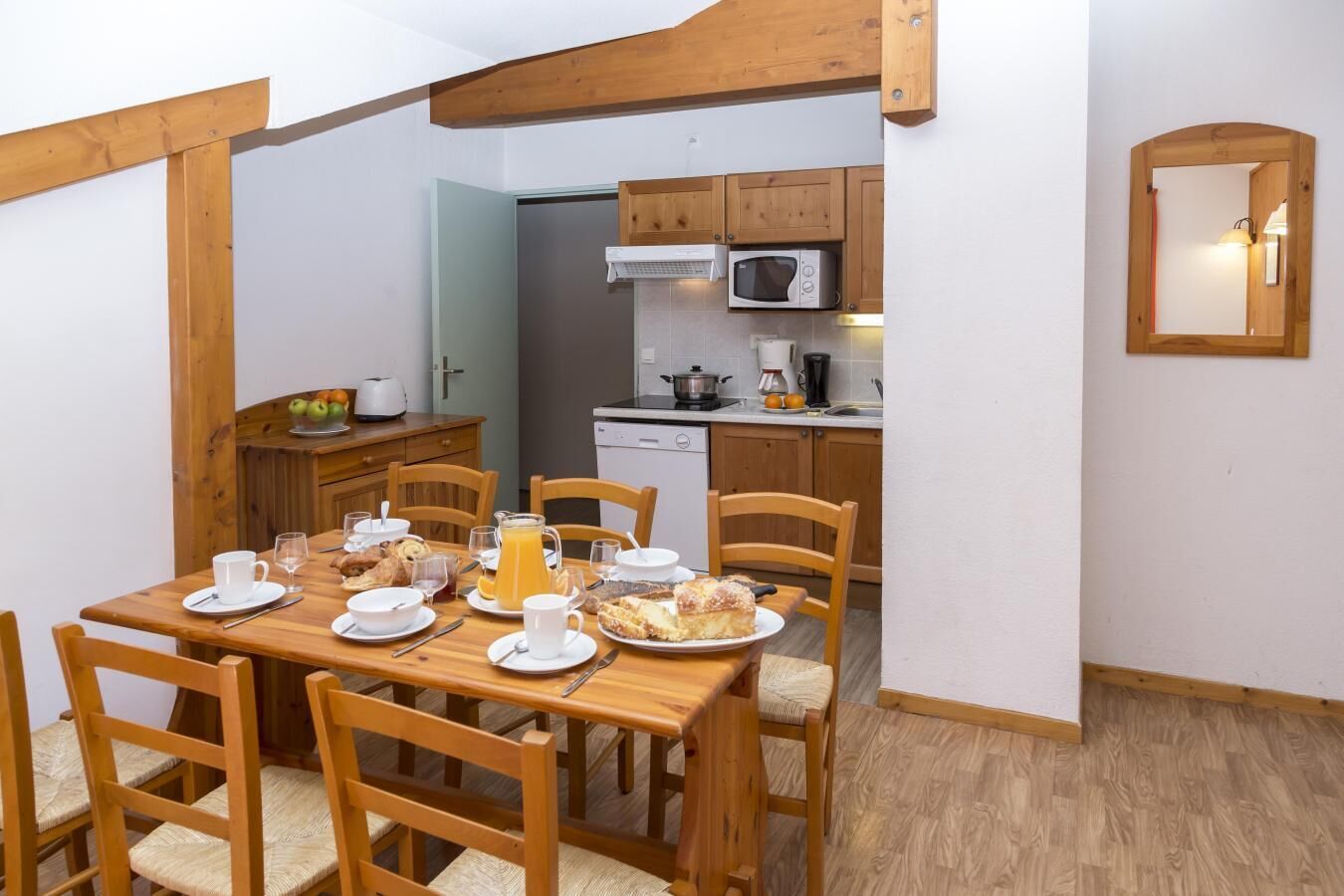 Un'area pranzo con un tavolo di legno apparecchiato per la colazione, inclusi piatti, tazze e una brocca di succo d'arancia. Sono presenti sedie di legno e un mobiletto in legno con un forno a microonde sopra il lavello nella zona cucina.