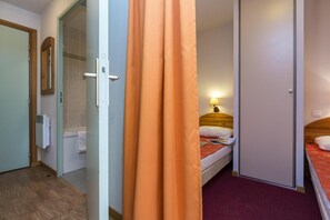 A hotel room with a wooden bed, a nightstand with a lamp, a bathroom with a shower, and a door with a curtain.