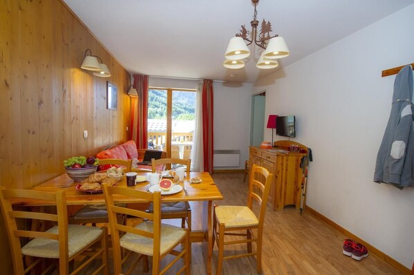 A cozy dining area with a wooden table, chairs, and a bowl of fruit. There is a sofa and a television in the background.