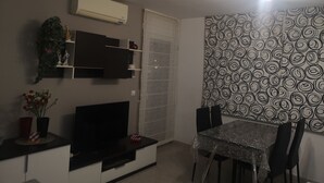 A living room with a flat-screen TV, a dining table with chairs, a patterned wall, and a ceiling light fixture.