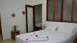 Una cama bien hecha con sábanas blancas y decoración floral rosa, una mesita de noche de madera con una lámpara, y una planta en maceta sobre una mesita auxiliar.