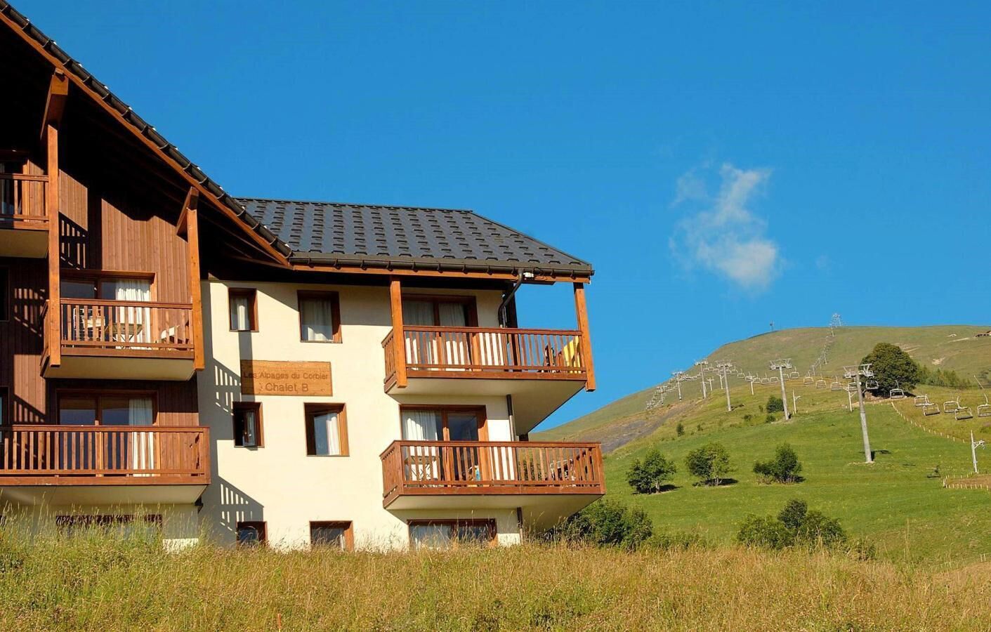 A multi-story building with balconies, a sign indicating 'Chalet B', and a view of a hilly landscape with ski lifts in the background.
