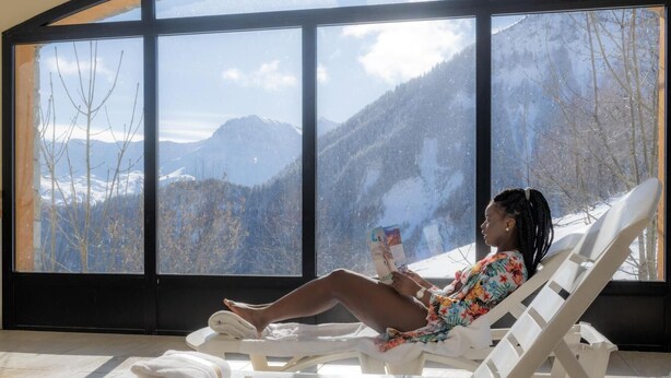 A person relaxing on a sun lounger with a book, in a room with large windows overlooking a snowy mountain landscape. - Ski-in/ski-out & foot of the slopes, fitness, swimming pool, sauna, parking, tv, 29-32m², Le Corbier (Le Corbier)