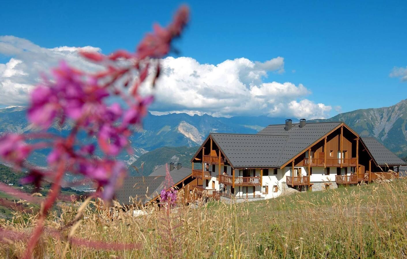 A wooden chalet with a sloped roof, multiple balconies, and a view of mountains in the background.