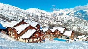 A cluster of wooden chalets with sloped roofs, set against a backdrop of snow-covered mountains under a clear blue sky. - Ski-in/ski-out & foot of the slopes, fitness, swimming pool, sauna, parking, tv, 29-32m², Le Corbier (Le Corbier)