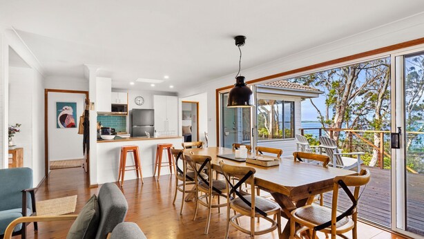 A modern kitchen with white cabinets, a black refrigerator, and a wooden dining table with chairs. There is a large window with a view of trees and a body of water.