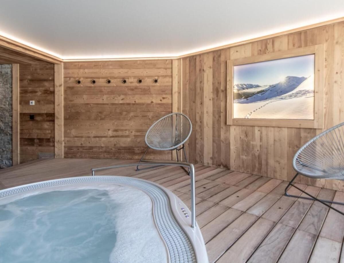 Ein Wellnessbereich mit Whirlpool, zwei Metallstühlen, Holzwänden und einem gerahmten Bild einer verschneiten Landschaft.