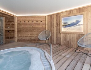 Un'area benessere con vasca idromassaggio, due sedie in metallo, pareti in legno e un quadro incorniciato raffigurante un paesaggio innevato.