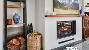 Un salon moderne avec une cheminée, des étagères en bois, un panier en osier et un tableau représentant un paysage de montagne.