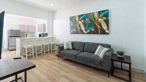 A modern living room with a grey sofa, a dining area with white chairs, a kitchen in the background, and a colorful abstract painting on the wall.