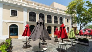 An outdoor patio with tables and red umbrellas, a building with arched windows, and a tree in the background.