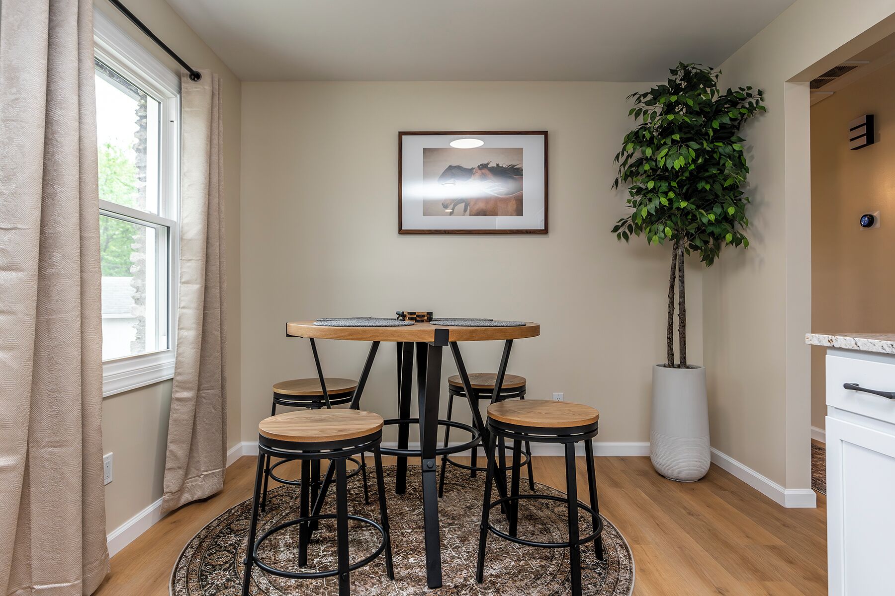 Un coin repas avec une table ronde et quatre tabourets, une grande plante en pot et un tableau encadré au mur.