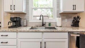 A kitchen with white cabinets, a Keurig coffee maker, a sink, and a window with a view of trees. - Charming Kentucky Stone House (Lexington)