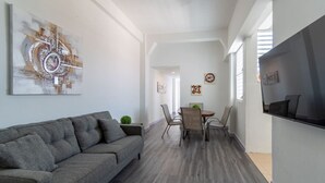 Ein modernes Wohnzimmer mit grauem Sofa, einem Essbereich mit Tisch und Stühlen sowie einem an der Wand befestigten Fernseher.