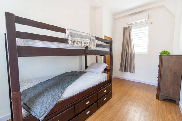 A bunk bed with a wooden frame, a gray blanket, and a pillow. There is a small wooden dresser to the right and a window with curtains.