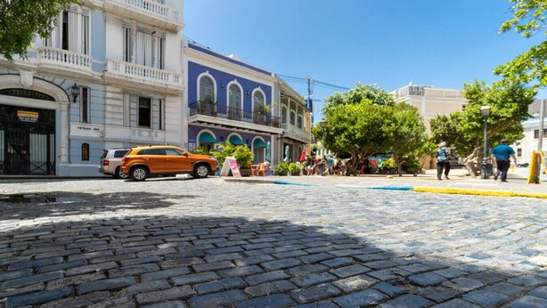 A cobblestone street with a parked orange car, historic buildings, and pedestrians. - Modern 2-BR Apt. in historic Old San Juan (San Juan)