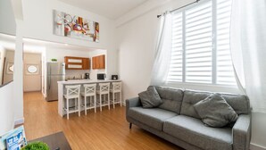 A modern living room with a grey sofa, wooden flooring, a kitchen in the background, and a large window with blinds. - Modern 2-BR Apt. in historic Old San Juan (San Juan)