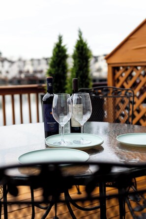 A table with two wine glasses and a bottle on a deck with a wooden railing and a view of buildings in the background.