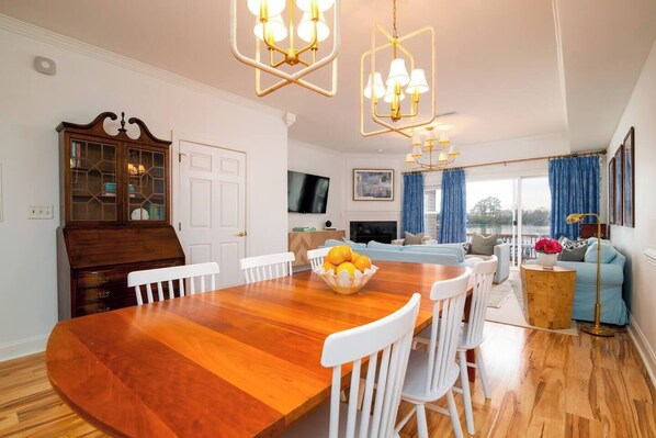A spacious living room with a wooden dining table, white chairs, a large sofa, and a TV mounted on the wall.