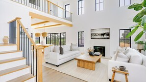 A spacious living room with a high ceiling, a wooden staircase, a fireplace, and large windows.