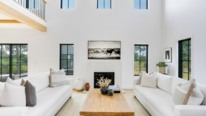 A spacious living room with a fireplace, large windows, and a high ceiling.