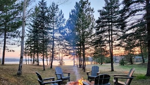 Un camping con cuatro sillas Adirondack dispuestas alrededor de una fogata, rodeado de altos pinos y vistas a un lago al atardecer.