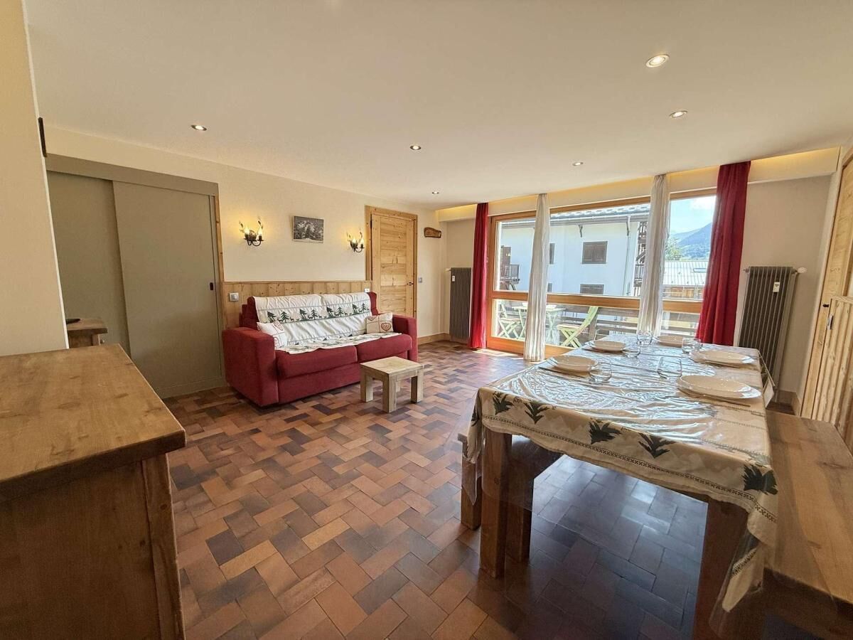 A spacious room with a herringbone floor, a red sofa, a wooden dining table set for four, and a large window with a view of a building and mountains.