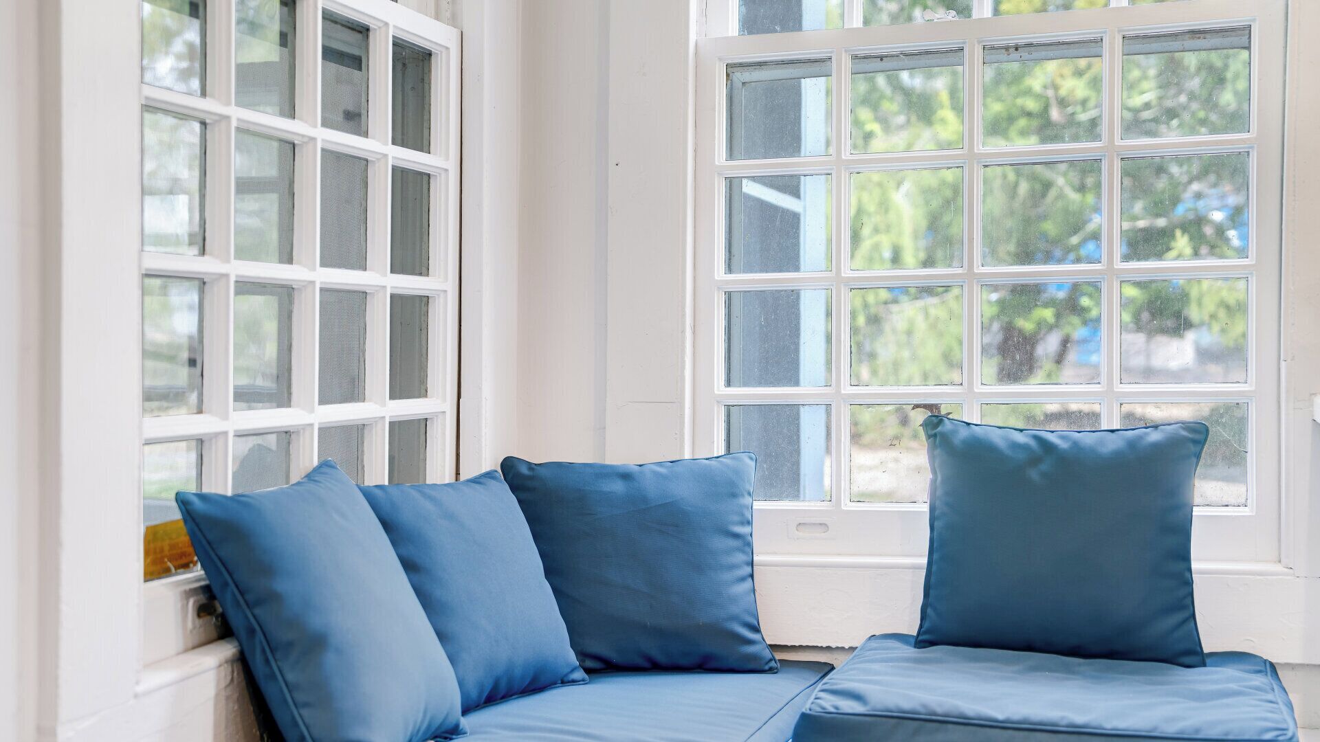 Una habitación con un ventanal blanco de múltiples paneles, un sofá azul con cojines a juego y vistas al exterior con vegetación.