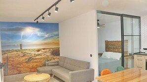 A modern living room with a large painting of a desert landscape, a grey sofa, a round wooden coffee table, and a glass dining table.