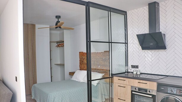A modern bedroom with a bed, a glass partition, a wardrobe, a ceiling fan, and a kitchen area with appliances.
