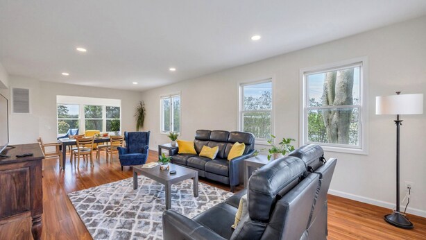 A spacious living room with a leather sofa, a dining area with wooden chairs, and large windows offering views of trees. - Cozy North Fork Home Among the Best Wineries in the Northeast, Minutes to... (Aquebogue)