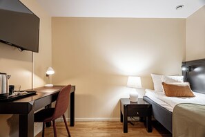 Quarto de hotel com uma cama, uma escrivaninha, uma cadeira, uma TV fixada na parede e um abajur.