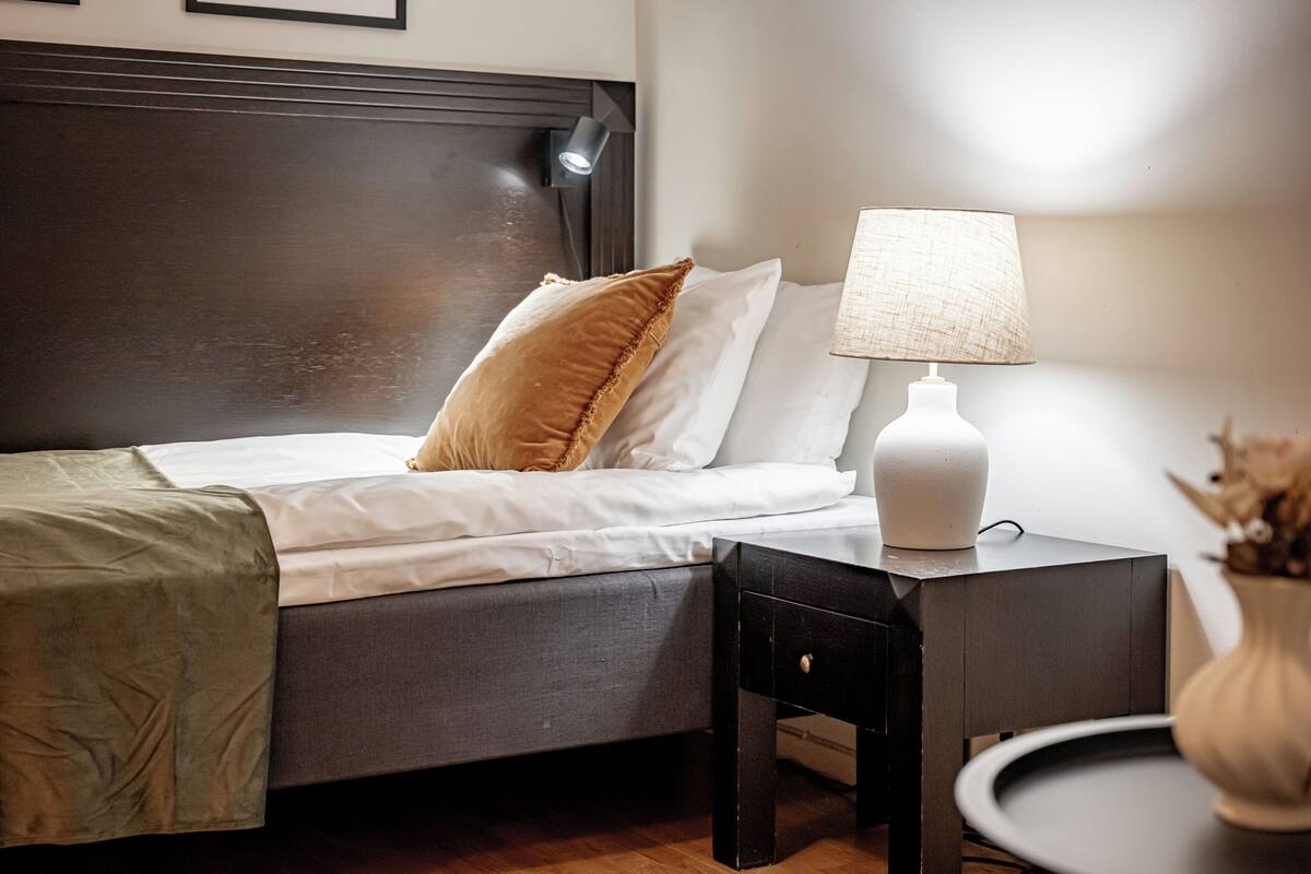 Un lit bien fait, avec une tête de lit sombre, du linge de lit blanc et un seul oreiller marron. Une lampe blanche, dotée d’un abat-jour texturé, se trouve sur une table de chevet, et un vase contenant des fleurs séchées est visible dans un coin.