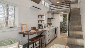 A modern kitchen with white cabinetry, a wooden countertop, and a built-in stove. There is a window with blinds, a staircase with a wooden railing, and a small seating area with stools.