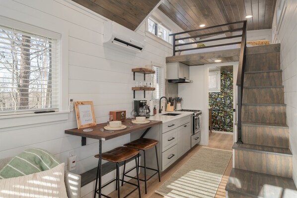 A modern kitchen with white cabinetry, a wooden countertop, and a built-in stove. There is a window with blinds, a staircase with a wooden railing, and a small seating area with stools.