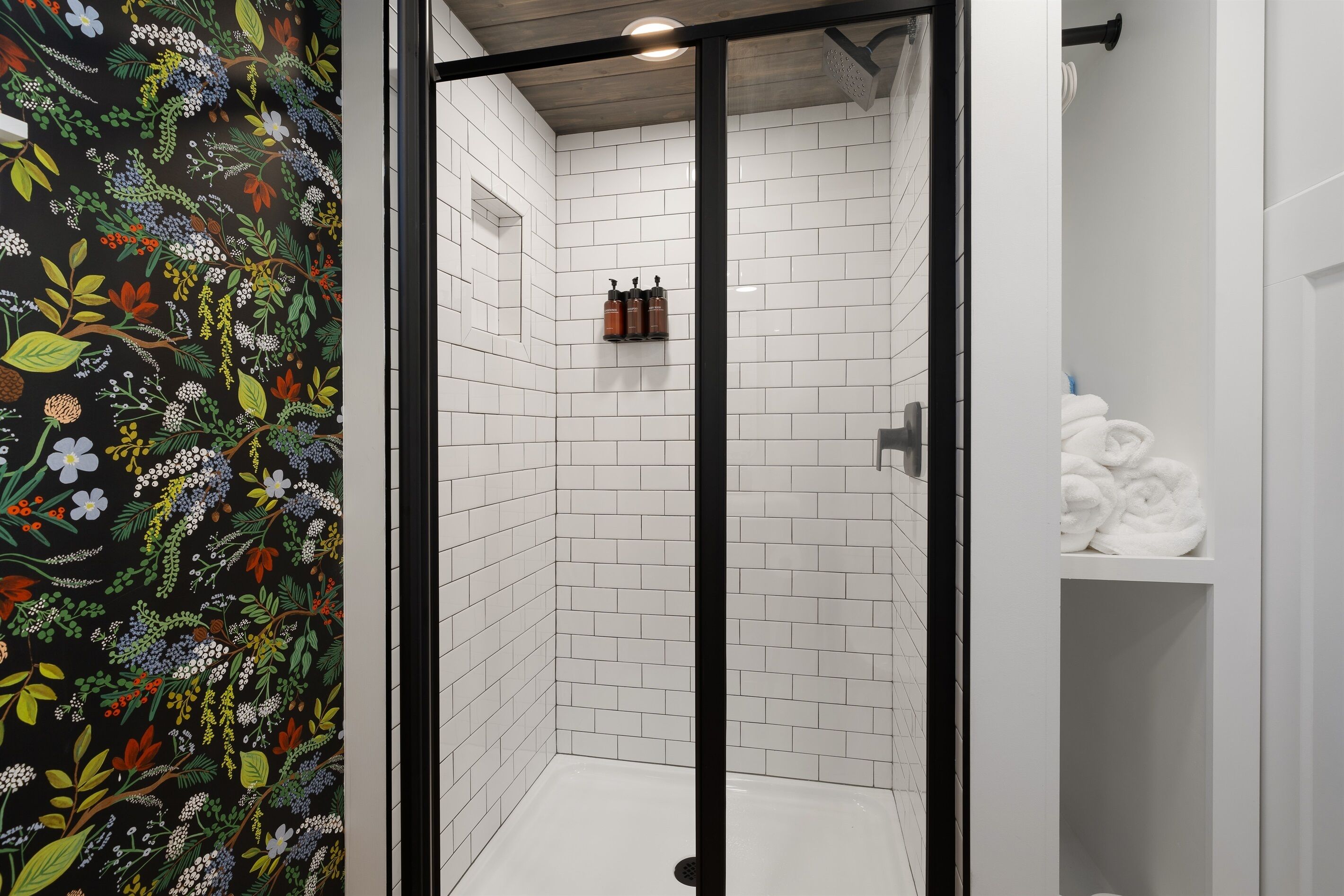 A shower area with white tiled walls, a glass door, and a black frame. There are bottles on a shelf above the shower and a stack of white towels to the right.