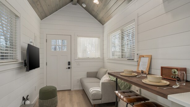A compact kitchen with a wooden ceiling, white walls, a grey sofa, a wooden bar counter, and a television mounted on the wall.