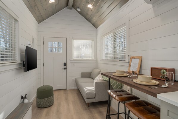A compact kitchen with a wooden ceiling, white walls, a grey sofa, a wooden bar counter, and a television mounted on the wall.