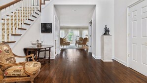 A spacious hallway with a wooden staircase, a dining area with a table and chairs, and a living room with a sofa and chairs.