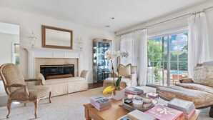A spacious living room with a fireplace, a large window with a view of greenery, and a collection of books and decorative items on a coffee table.