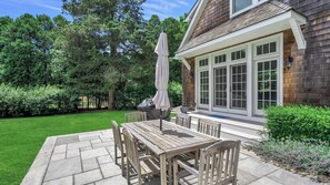 Relax in the serene outdoor patio area with a wooden table and chairs, surrounded by lush greenery and a beautifully maintained lawn.