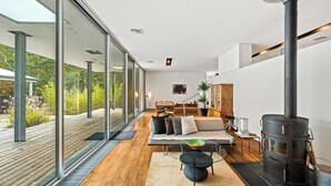 A modern living room with a large glass wall, a wooden deck, a sofa, a dining table, and a wood-burning stove. - Modern home sited on a lush wood acre, stunning pool, covered deck, outdoor... (East Hampton)
